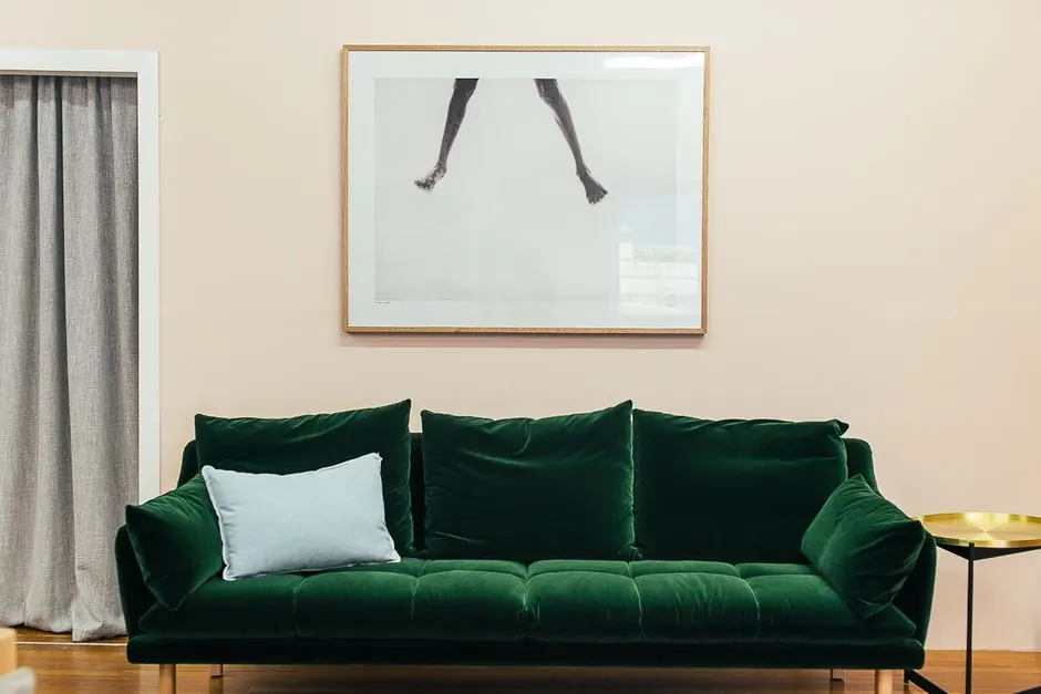 Comfortable green velour sofa with soft cushions and small round table placed on parquet floor near wall with creative framed picture in stylish living room